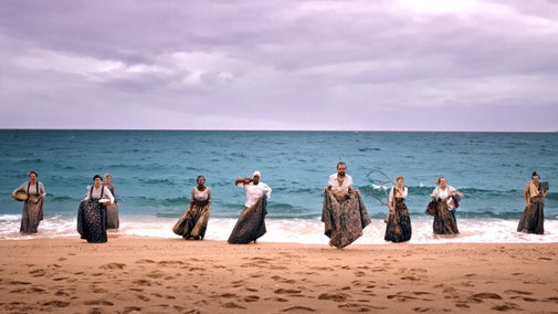 Nine performers in long skirts walking out of the sea onto a sandy beach